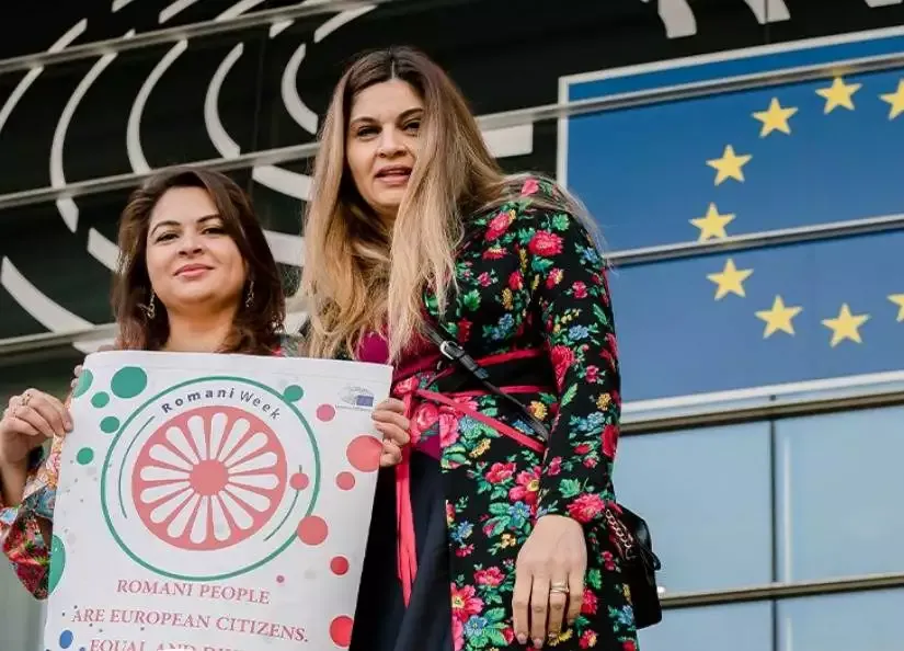 Two people holding Romani Week sign outside building.